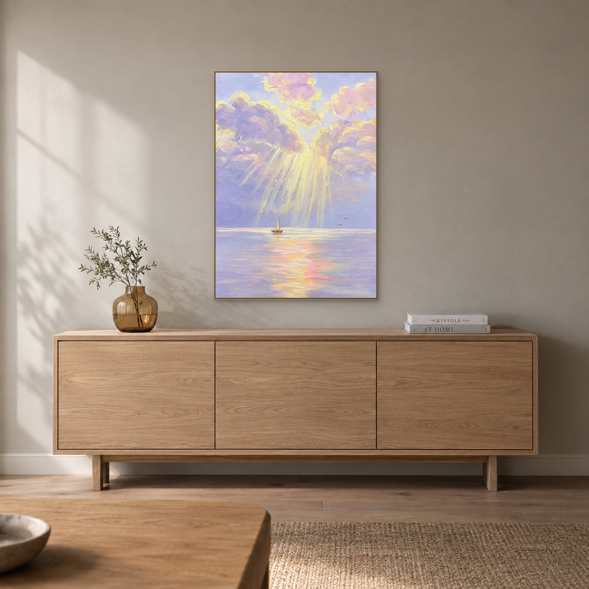 Paysage marin pastel dans un cadre champagne, au-dessus d’un buffet en bois clair avec un petit vase ambré et des livres.