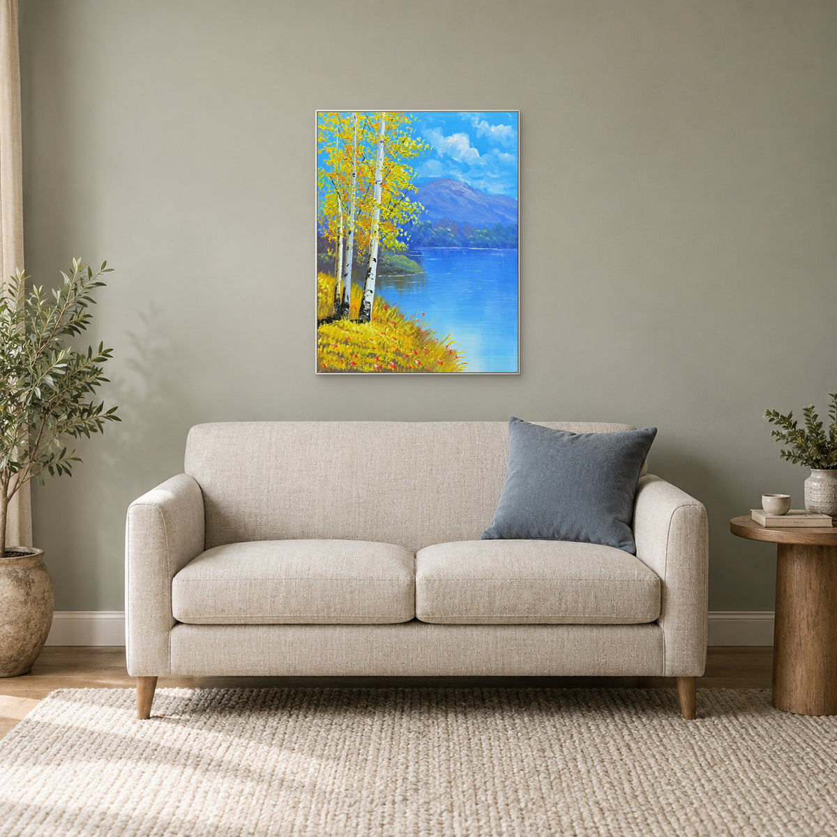 Paysage de bouleaux et de lac dans un cadre blanc au-dessus d’un petit canapé crème, avec coussin bleu et plantes.