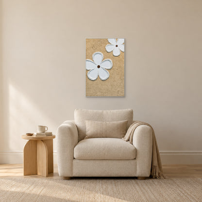Deux fleurs blanches sur fond beige, cadre blanc, au-dessus d’un fauteuil crème près d’une petite table ton chêne