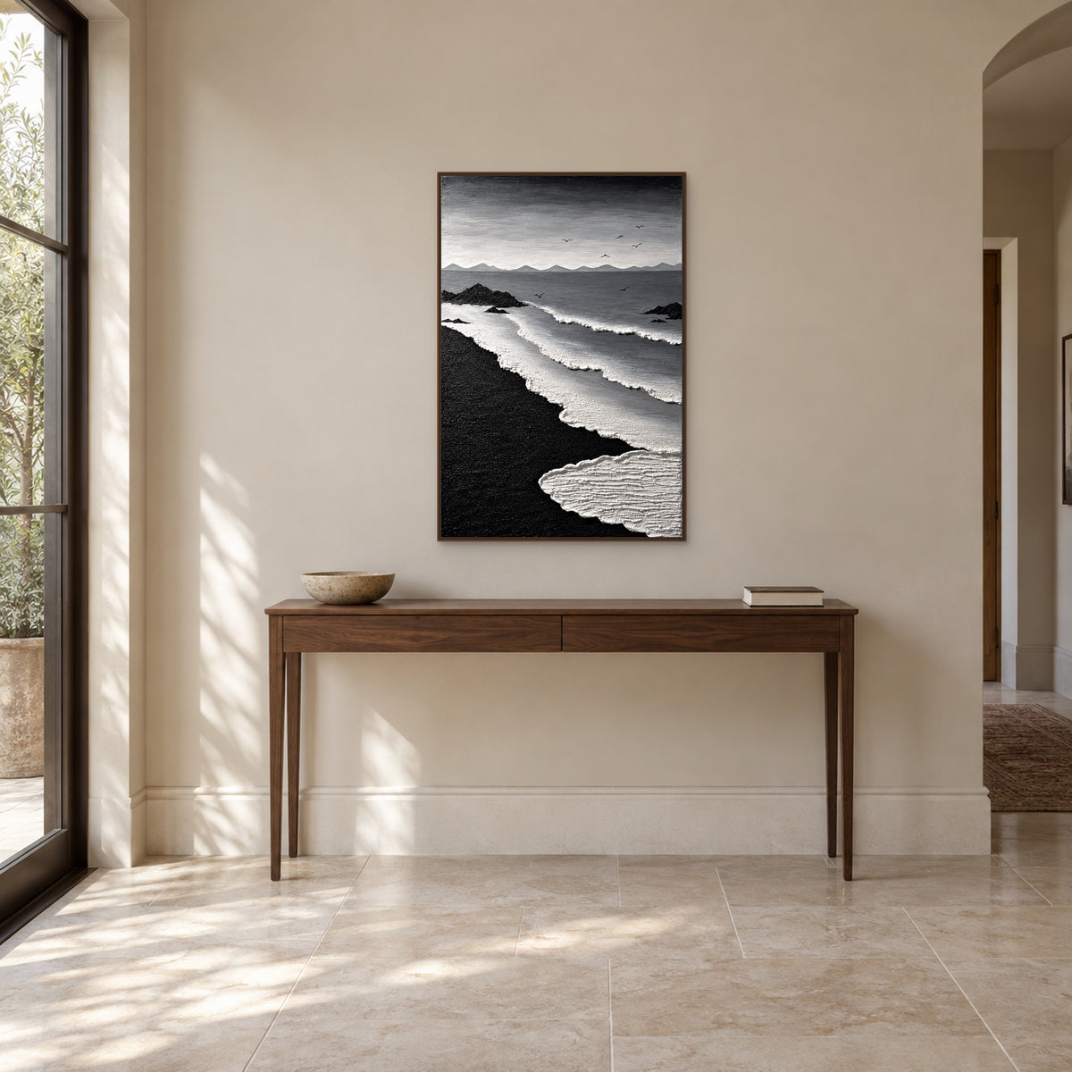 Tableau de sable noir et d’écume blanche dans un cadre flottant ton noyer, au-dessus d’une fine console d’entrée avec un bol bas et des livres